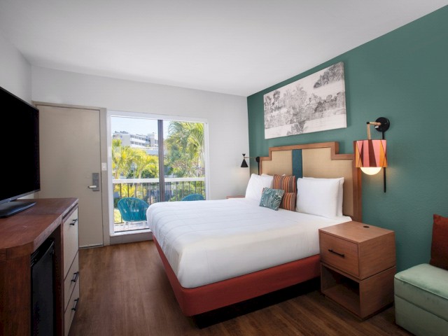 Modern hotel room with a large bed, TV, artwork, and a view to the outdoors.