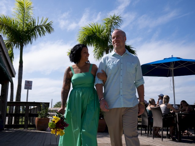Two people walking hand-in-hand, dressed for a special occasion, with palm trees and a sunny sky.