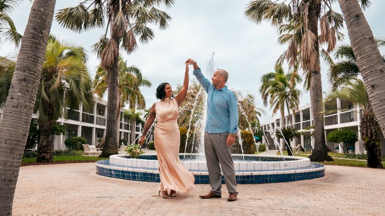 Two people are dancing by a fountain surrounded by palm trees.