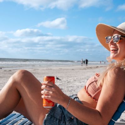 Woman relaxing on a beach chair with a drink, wearing sunglasses and a sunhat.