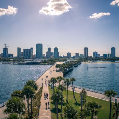 A sunny boardwalk with people, overlooking a bay with a city skyline in the background.