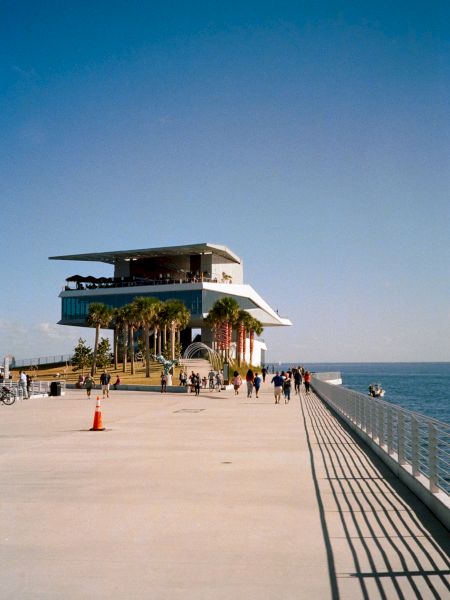 Sunny pier with people, railing, ocean view, and a structure at the end.