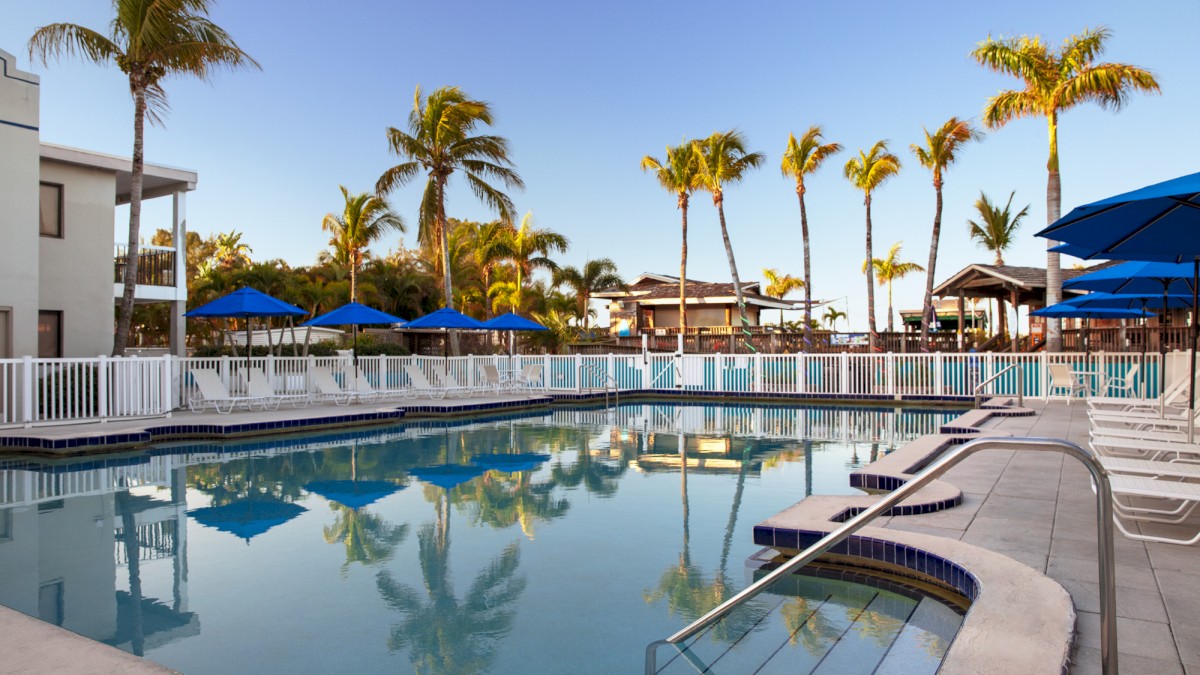 A large serene pool with umbrellas and palm trees reflecting in the water, under a clear sky.