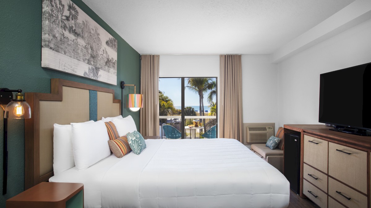 A modern hotel room with a bed, side tables, lamps, and a TV against a window backdrop.