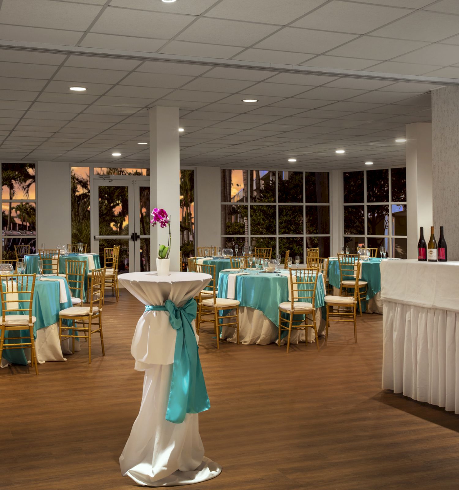 The image shows an event space with round tables covered in turquoise and white linens, high tables with decorations, and some wine bottles.