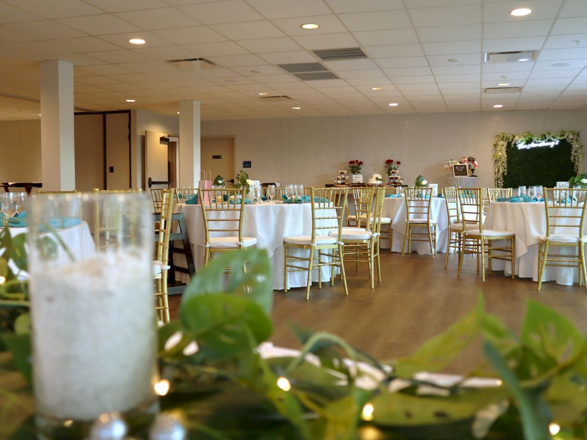 The image shows a decorated event hall with round tables, white tablecloths, gold chairs, and green foliage centerpieces, suggesting a celebration.
