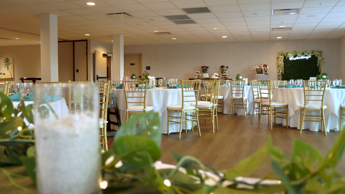 The image shows a decorated event hall with round tables, white tablecloths, gold chairs, and green foliage centerpieces, suggesting a celebration.