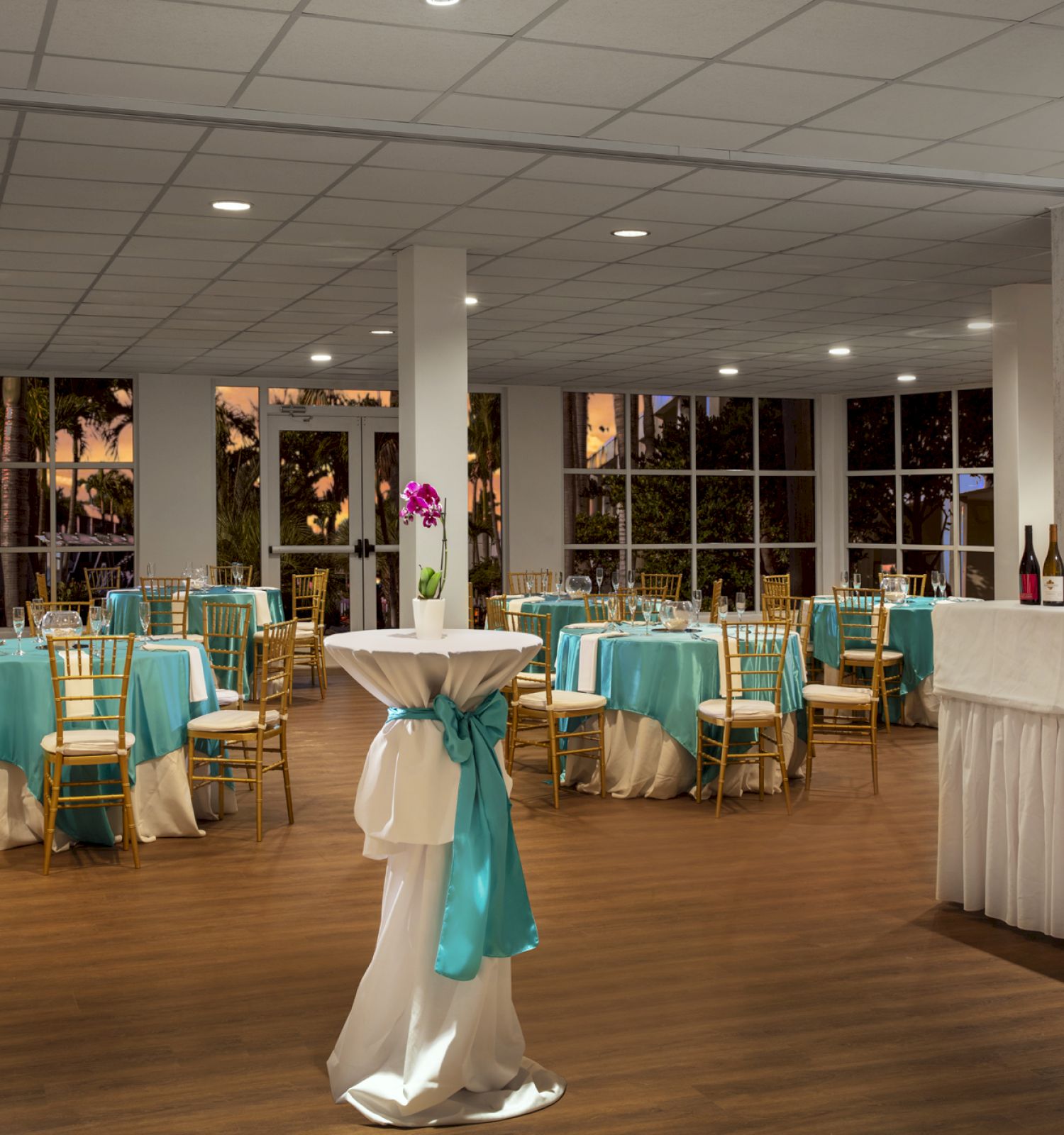 A decorated banquet hall with tables and chairs adorned in teal and white, high-top tables with flowers, and a bar area with bottles.