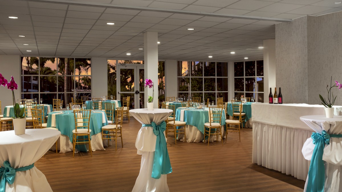 A decorated banquet hall with tables and chairs adorned in teal and white, high-top tables with flowers, and a bar area with bottles.