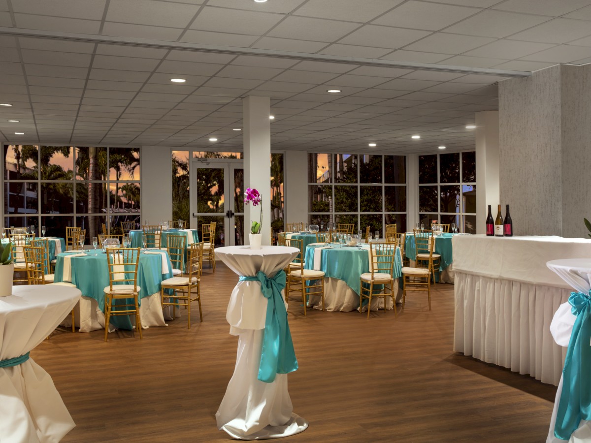 A decorated banquet hall with tables and chairs adorned in teal and white, high-top tables with flowers, and a bar area with bottles.