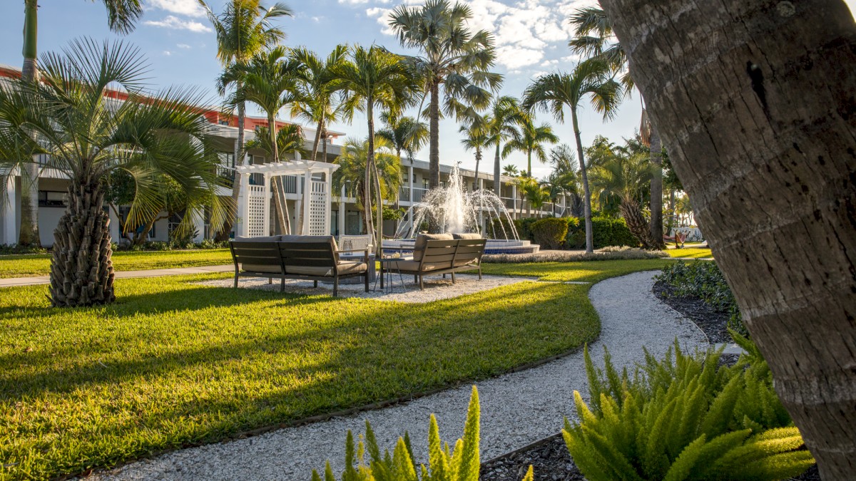 A lush courtyard garden with palm trees, a water fountain, and outdoor seating along a curved pathway, set against a backdrop of a building and blue skies.