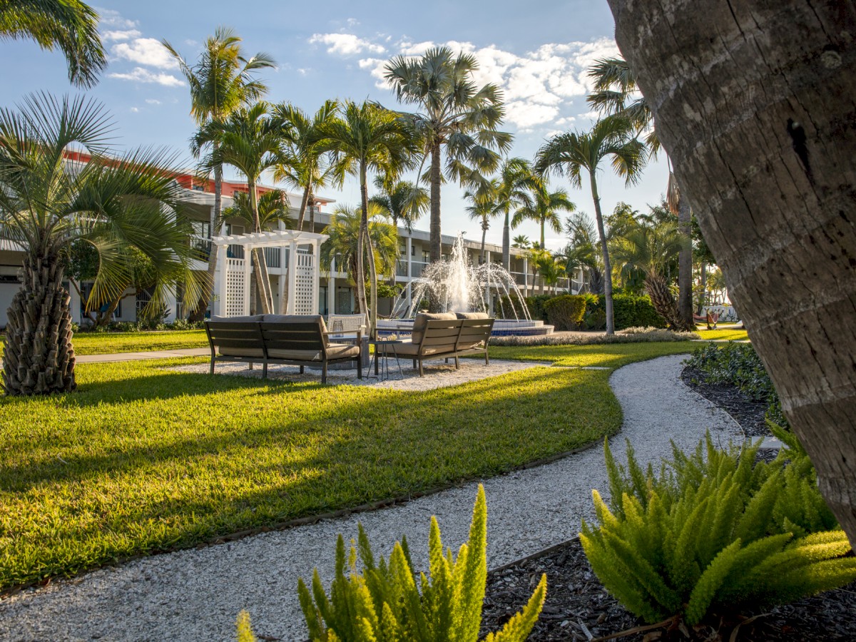 A lush courtyard garden with palm trees, a water fountain, and outdoor seating along a curved pathway, set against a backdrop of a building and blue skies.