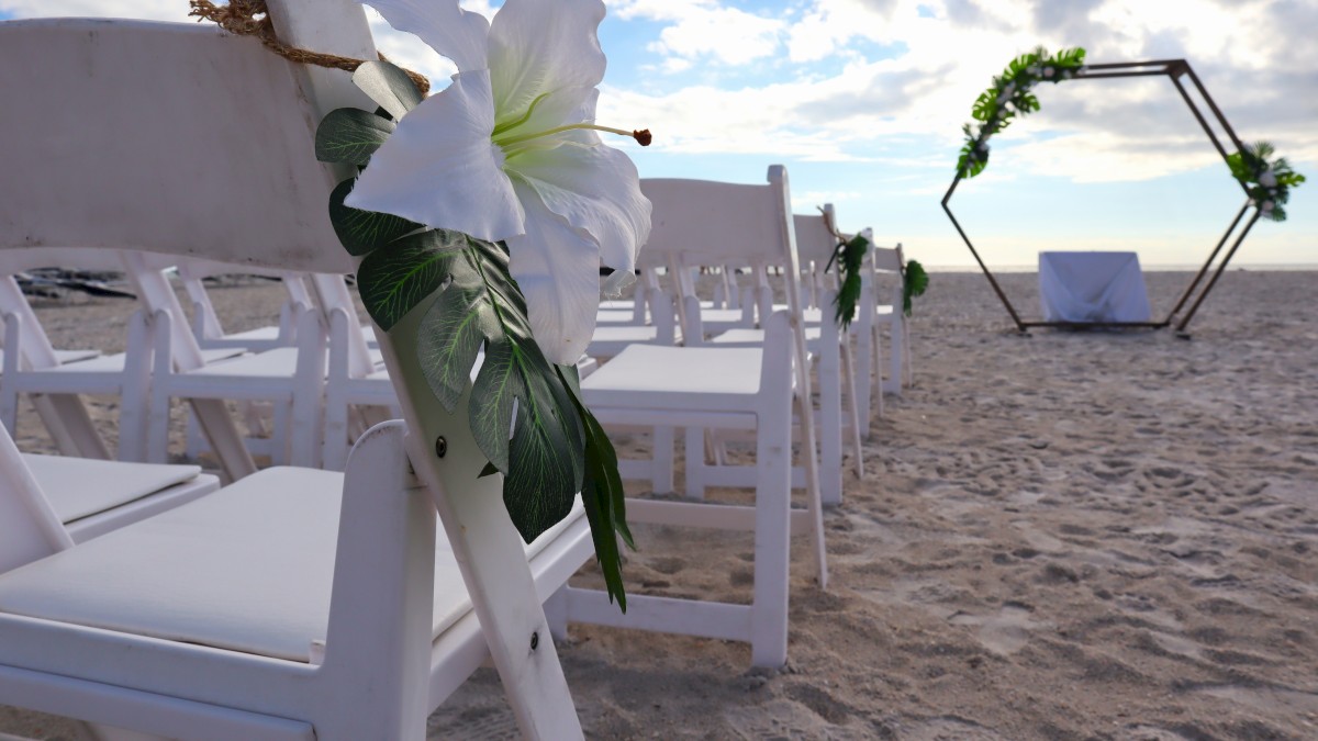 Outdoor beach wedding setup with white chairs, large white flowers, and a hexagonal arch decorated with greenery in the background, ending the sentence.