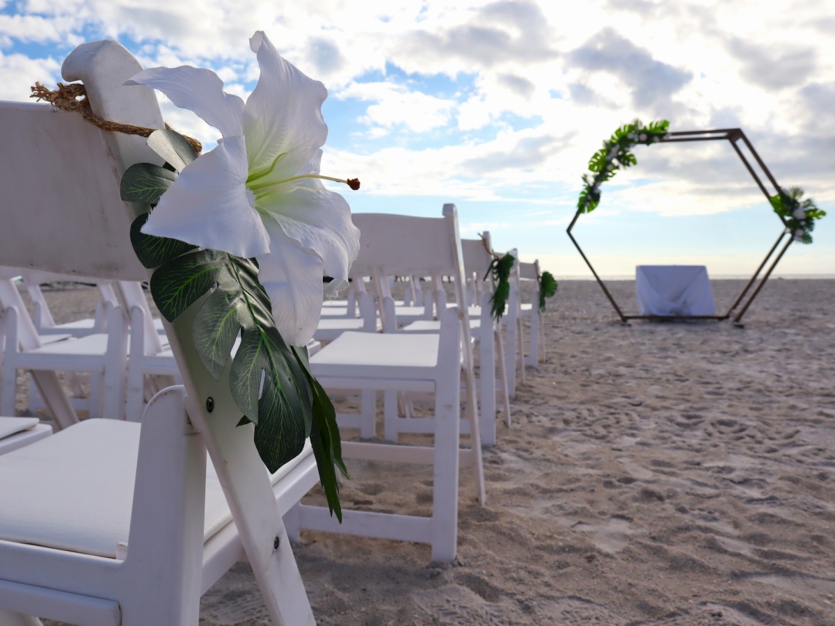 Outdoor beach wedding setup with white chairs, large white flowers, and a hexagonal arch decorated with greenery in the background, ending the sentence.