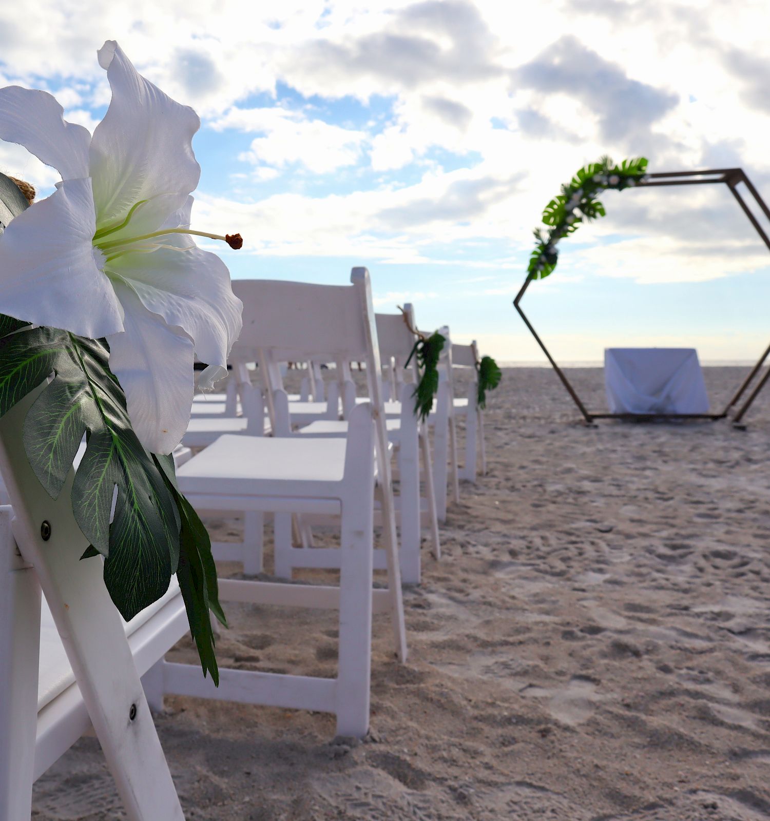 Outdoor beach wedding setup with white chairs, large white flowers, and a hexagonal arch decorated with greenery in the background, ending the sentence.