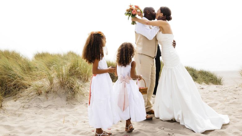 A couple is kissing on a beach, with the woman in a wedding dress, man in a tan suit, and two flower girls watching them.