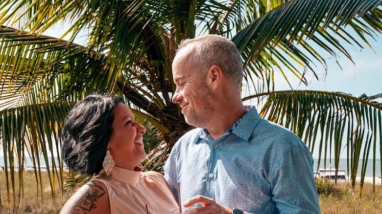 A couple is standing on a wooden deck near a palm tree, enjoying drinks and smiling at each other under a sunny sky.