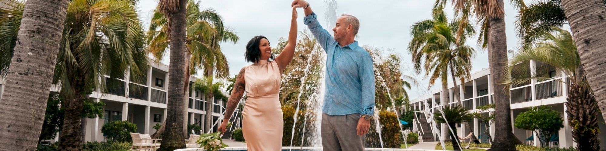 A couple dances in front of a fountain surrounded by palm trees and a building in the background, both smiling and holding hands, ending the sentence.
