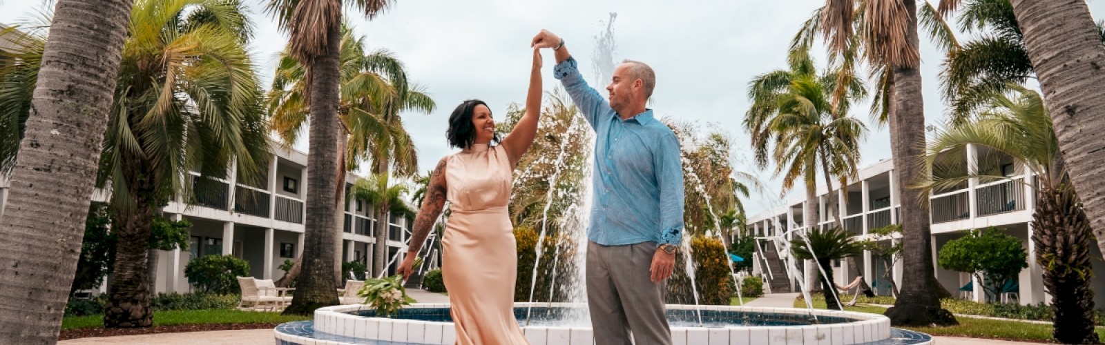 A couple dances in front of a fountain surrounded by palm trees and a building in the background, both smiling and holding hands, ending the sentence.