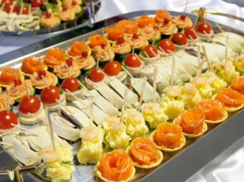 The image shows a platter of assorted appetizers, including canapés, salmon rolls, cheese, cherry tomatoes on skewers, and other bite-sized snacks.