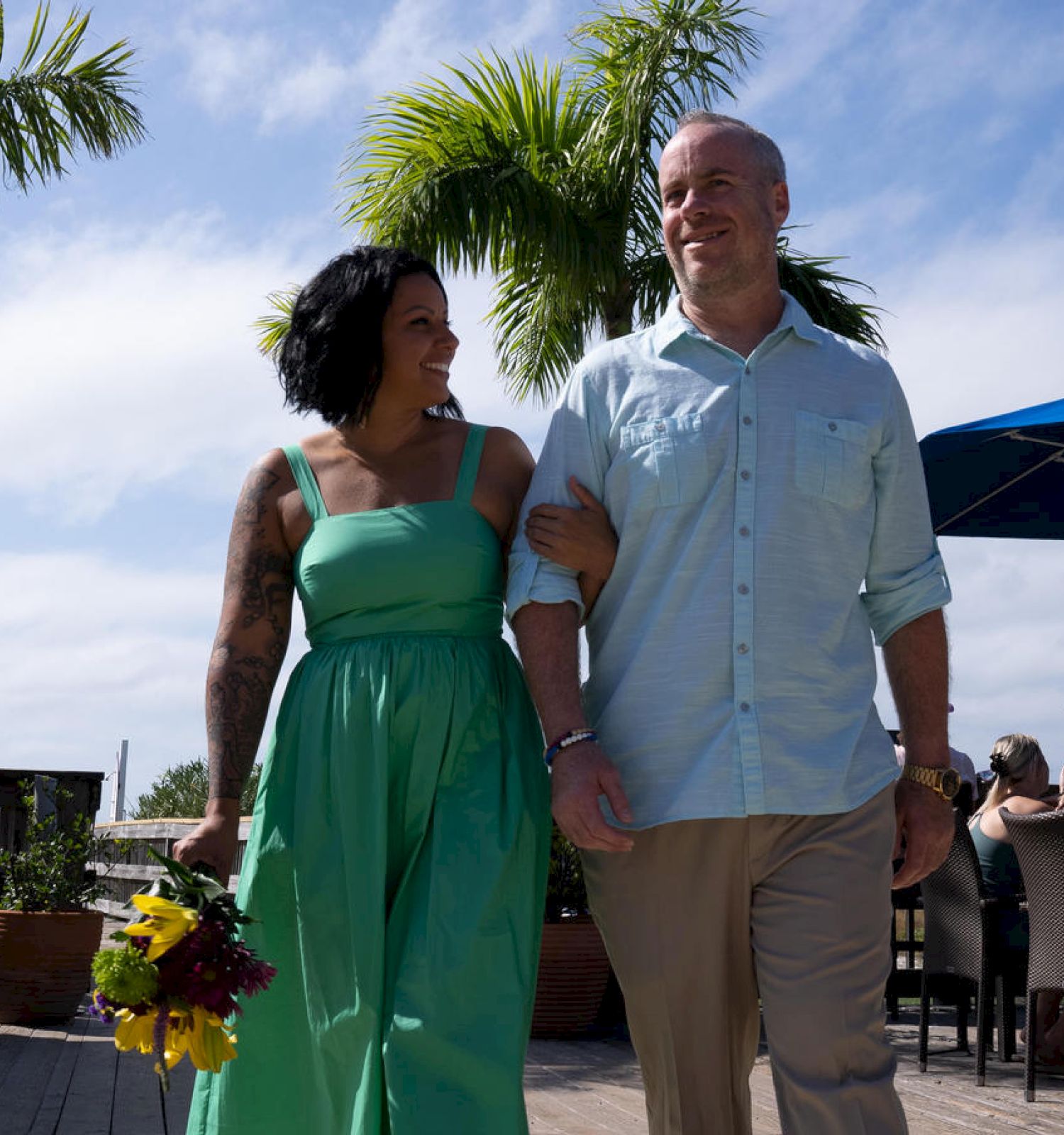A happy couple walks arm in arm under a sunny sky, with palm trees and an outdoor seating area in the background, the woman holding flowers.