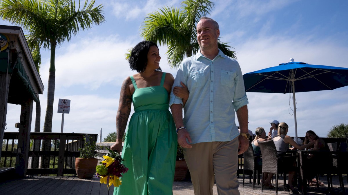 A happy couple walks arm in arm under a sunny sky, with palm trees and an outdoor seating area in the background, the woman holding flowers.