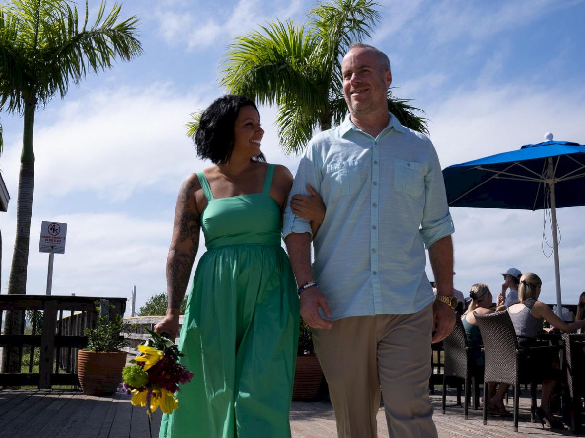 A happy couple walks arm in arm under a sunny sky, with palm trees and an outdoor seating area in the background, the woman holding flowers.