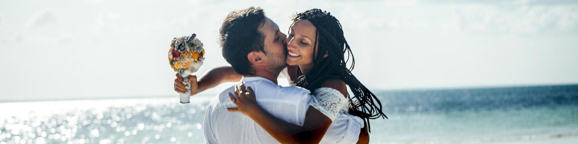 A couple is joyfully embracing on a beach; one person holding a bouquet, and the scene is set against a backdrop of the sea and the sky.