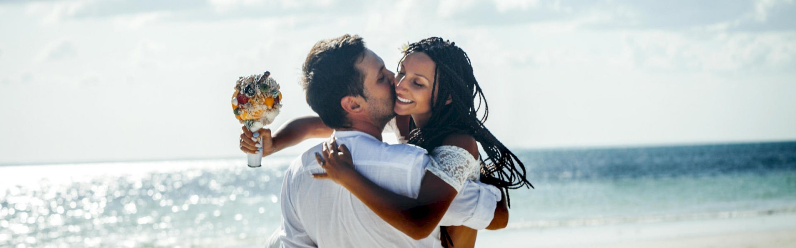 A couple is joyfully embracing on a beach; one person holding a bouquet, and the scene is set against a backdrop of the sea and the sky.