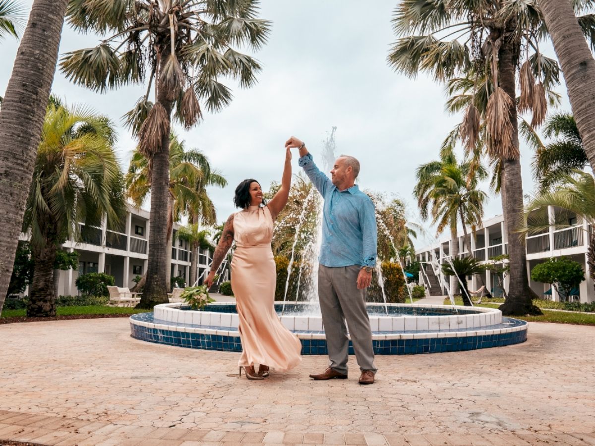 A couple dances near a fountain surrounded by palm trees, set against a backdrop of a tropical resort with buildings in the distance.