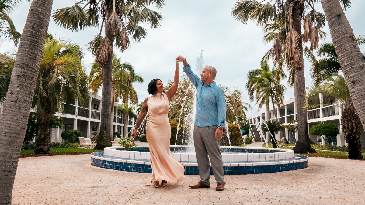 A couple dances near a fountain surrounded by palm trees, set against a backdrop of a tropical resort with buildings in the distance.