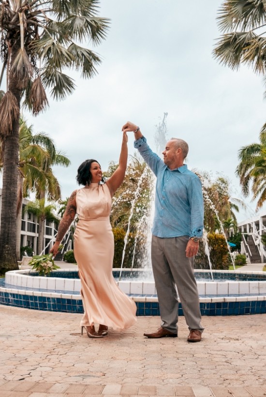 A couple dances near a fountain surrounded by palm trees, set against a backdrop of a tropical resort with buildings in the distance.