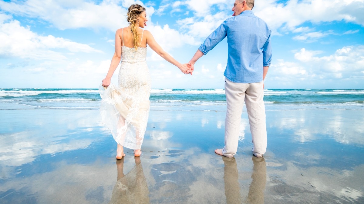 A couple holding hands stands on a beach with blue sky and clouds reflected in the wet sand. They face the ocean, creating a serene scene.