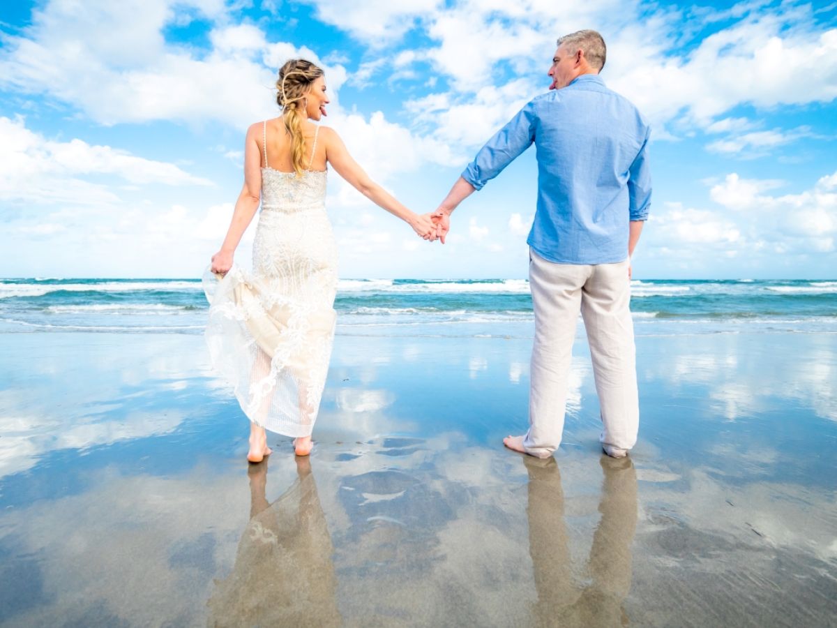 A couple holding hands stands on a beach with blue sky and clouds reflected in the wet sand. They face the ocean, creating a serene scene.