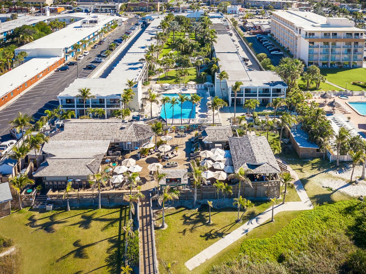 The image shows an aerial view of a resort with multiple buildings, pools, and outdoor seating areas surrounded by palm trees.