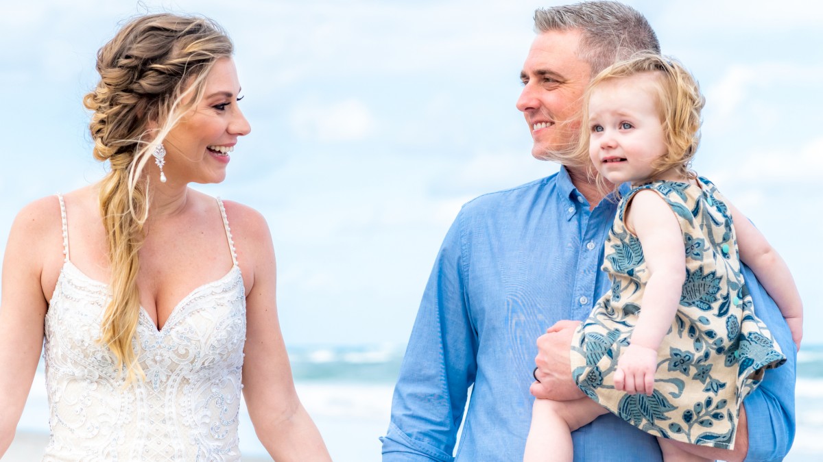 A woman in a white dress, a man holding a child, all smiling and standing on a beach under a blue sky.