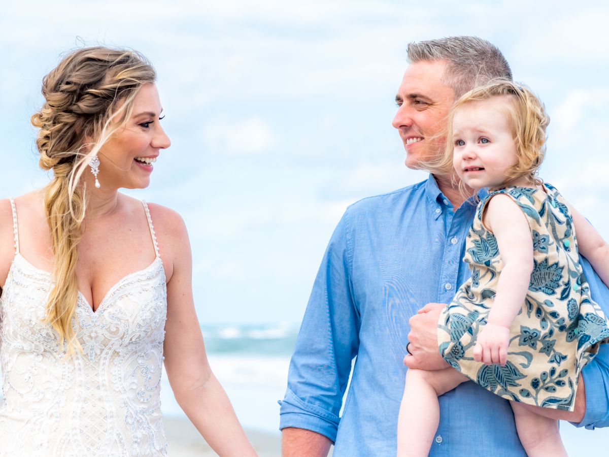 A woman in a white dress, a man holding a child, all smiling and standing on a beach under a blue sky.