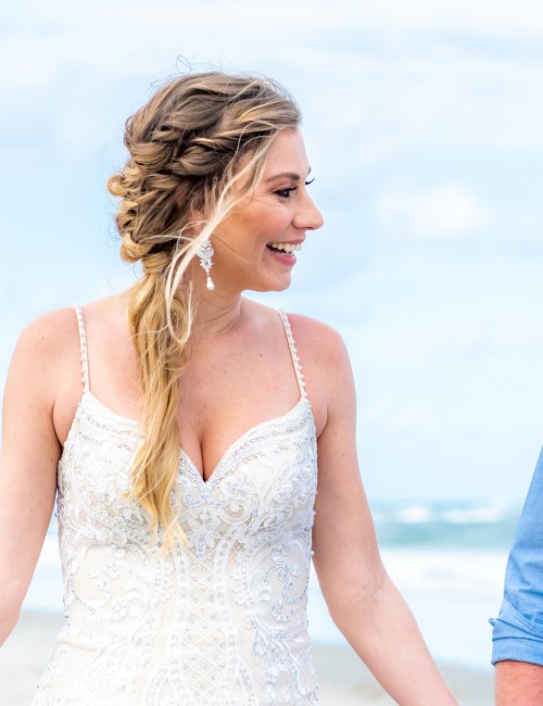 A woman in a white dress, a man holding a child, all smiling and standing on a beach under a blue sky.