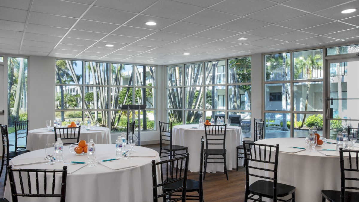 The image shows a bright, modern conference room with round tables covered in white tablecloths and black chairs, set up beside large windows.