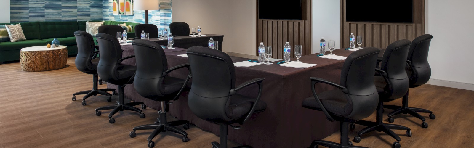 The image shows a modern conference room with a long table, chairs, bottled water, notepads, and two large screens on the wall in a well-lit space.