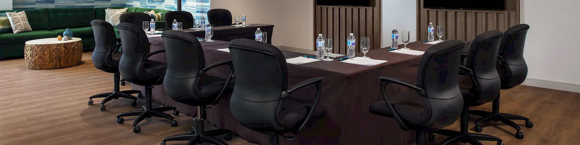 The image shows a modern conference room with a long table, chairs, bottled water, notepads, and two large screens on the wall in a well-lit space.