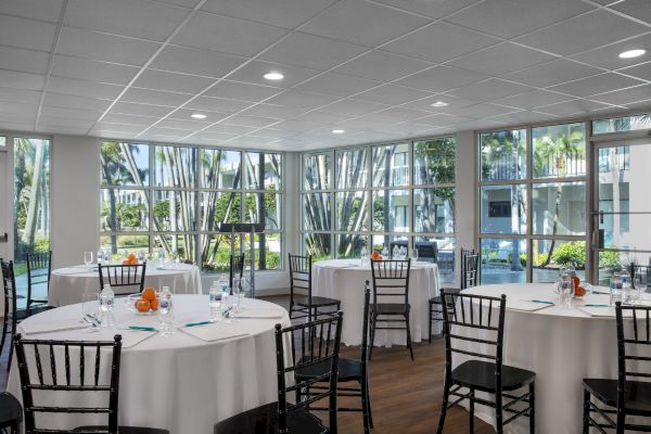 A bright room with round tables, white tablecloths, black chairs, and a wall of windows showing a green outdoor area with palm trees.