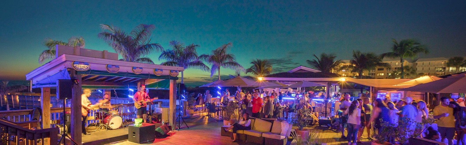 A lively outdoor scene at sunset features a band performing on a stage, palm trees, and people socializing under illuminated tents.