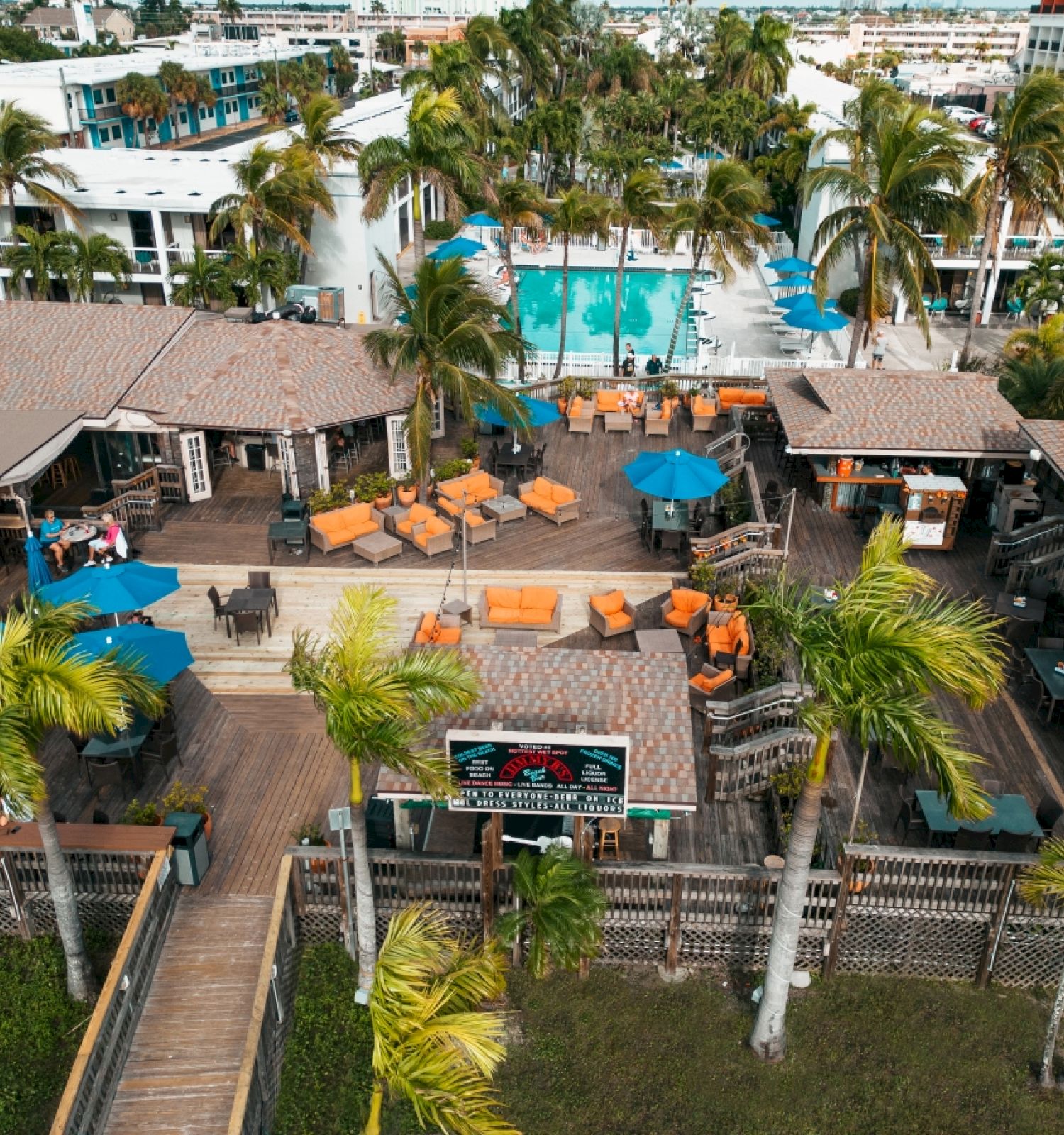 An outdoor beachside resort features a pool, numerous seating areas, umbrellas, palm trees, and a relaxed tropical atmosphere, ending the sentence.