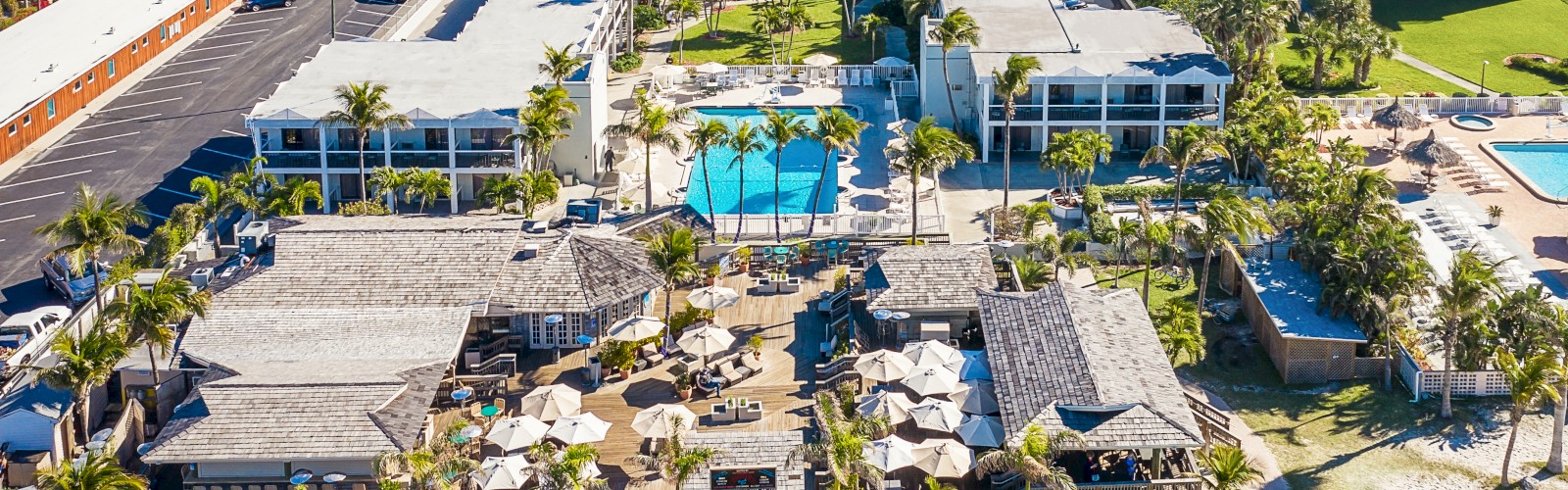 The image shows an aerial view of a resort with multiple buildings, swimming pools, palm trees, and pathways leading to beach huts and lounging areas.
