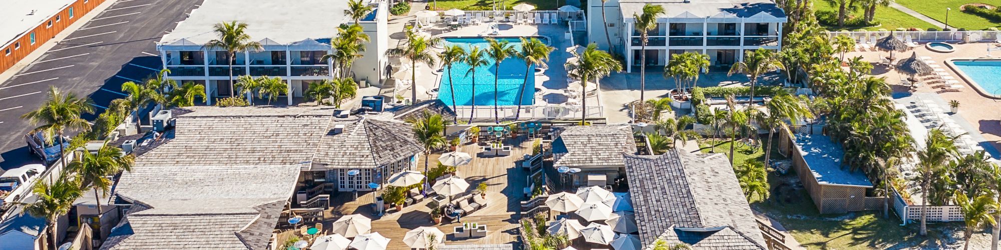 The image shows an aerial view of a resort with multiple buildings, swimming pools, palm trees, and pathways leading to beach huts and lounging areas.