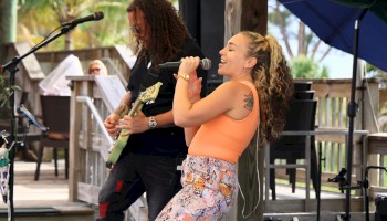 Two musicians perform on a sunny outdoor stage: a guitarist in the background and a woman in an orange top singing into a microphone, captivating the crowd.