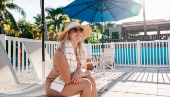 A smiling woman in a hat sits by a pool on a sunny day, with umbrellas and palm trees in the background.