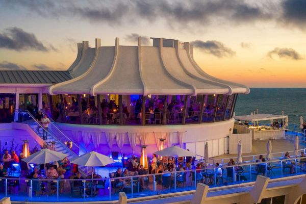 Cruise ship deck at sunset, with people, outdoor seating, and a vibrant sky.