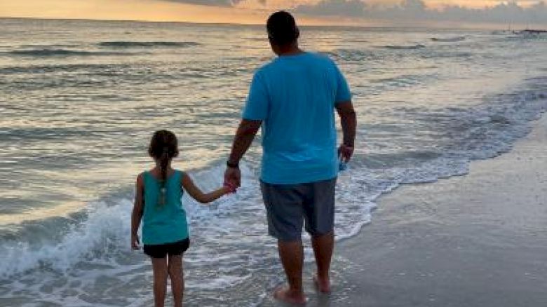 An adult and child walk on the beach, holding hands at sunset.